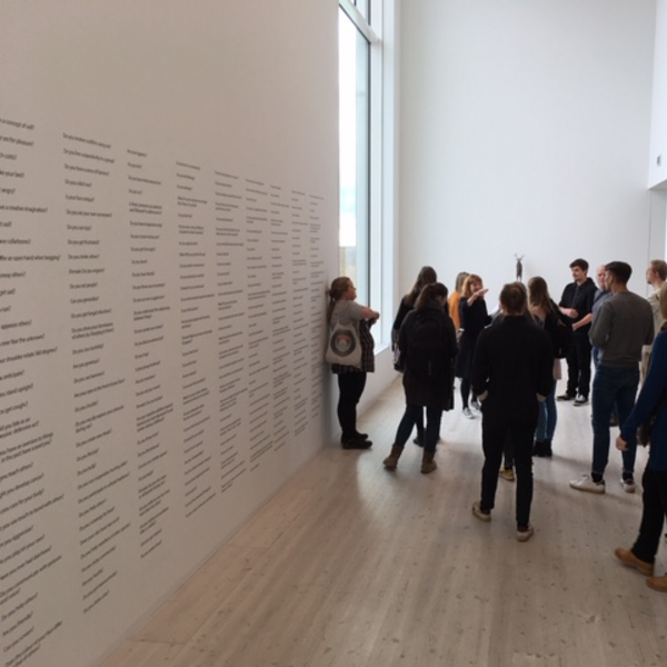 A group of students in a gallery in front of a long wall with multiple lists of content on it. 
