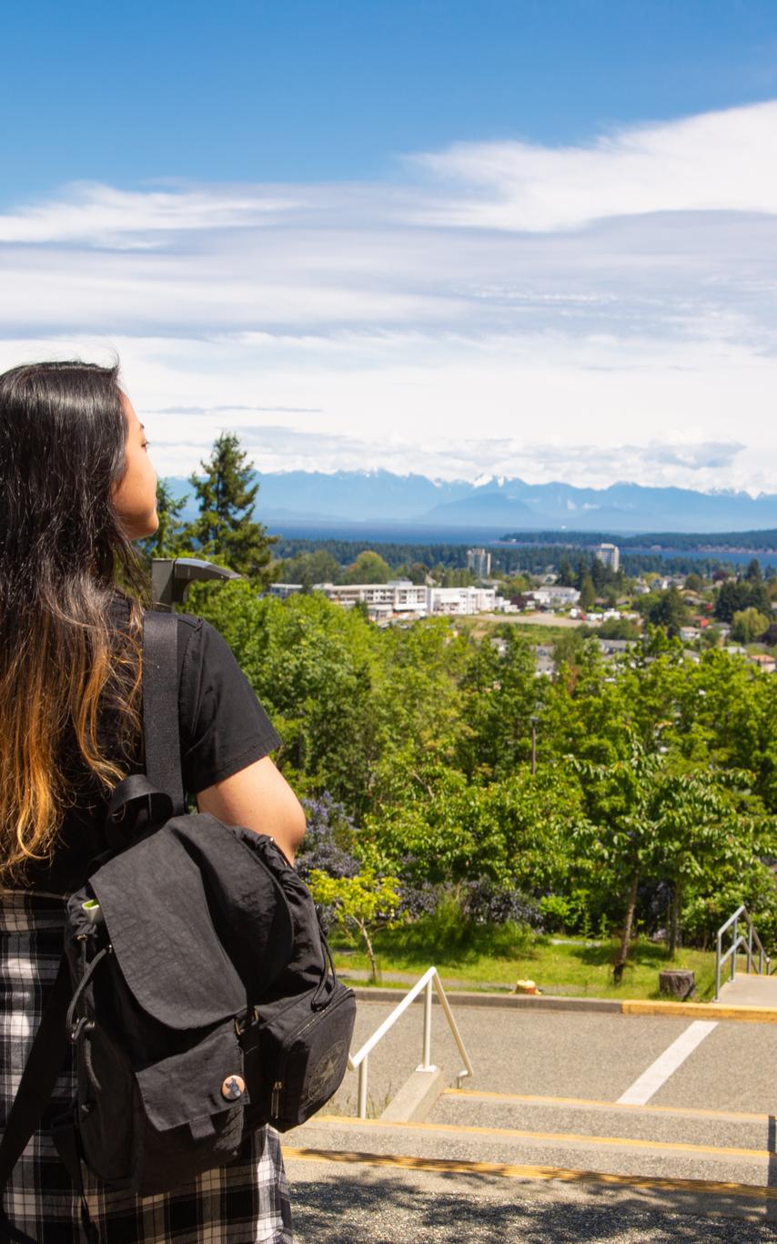 A young-adult standing at the top of stairs with long dark hair and a black backpack and a plaid shirt tied around her waist looking out at the trees and city in the distance.