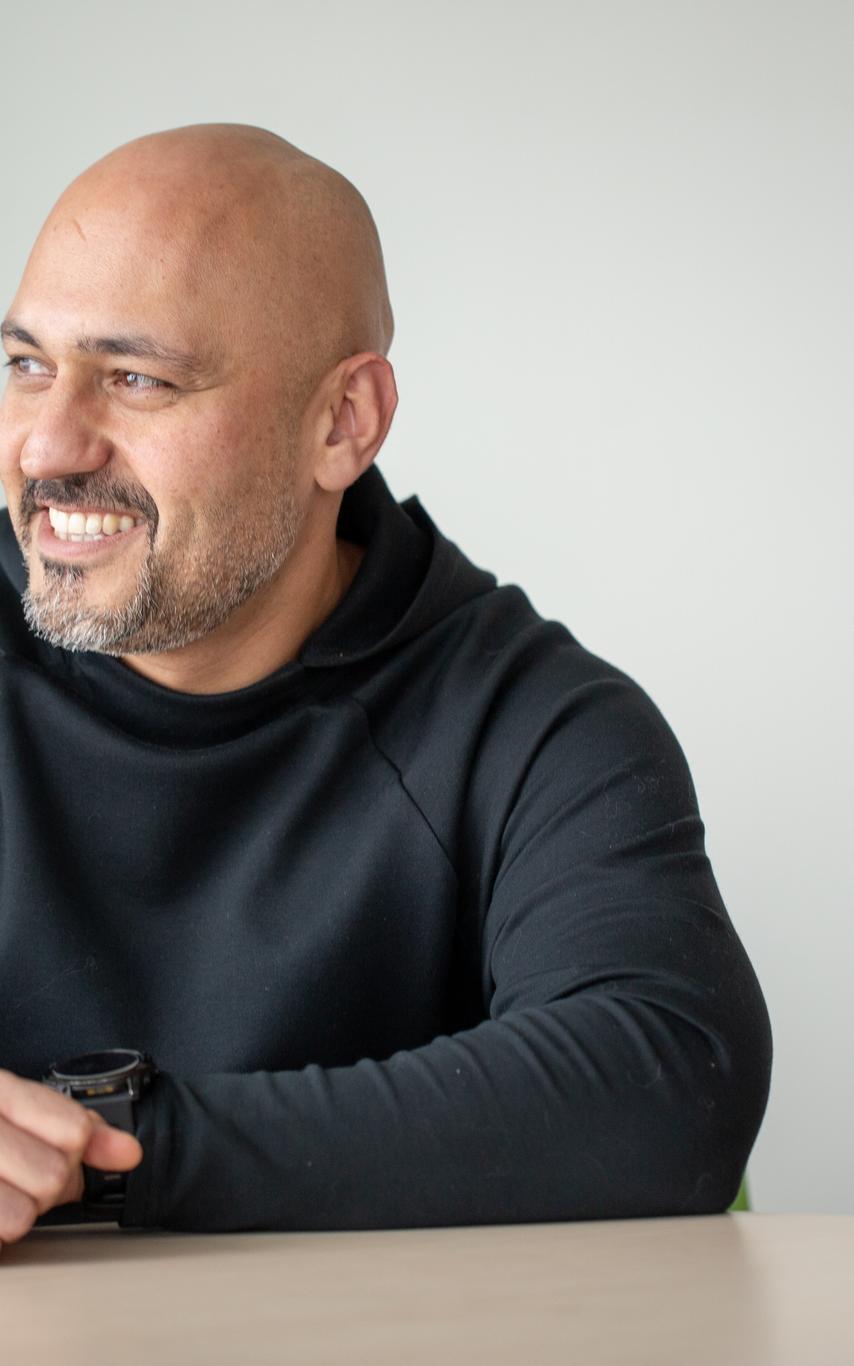 a bald man with a beard and mustache in a black sweater leaning on a table and smiling.