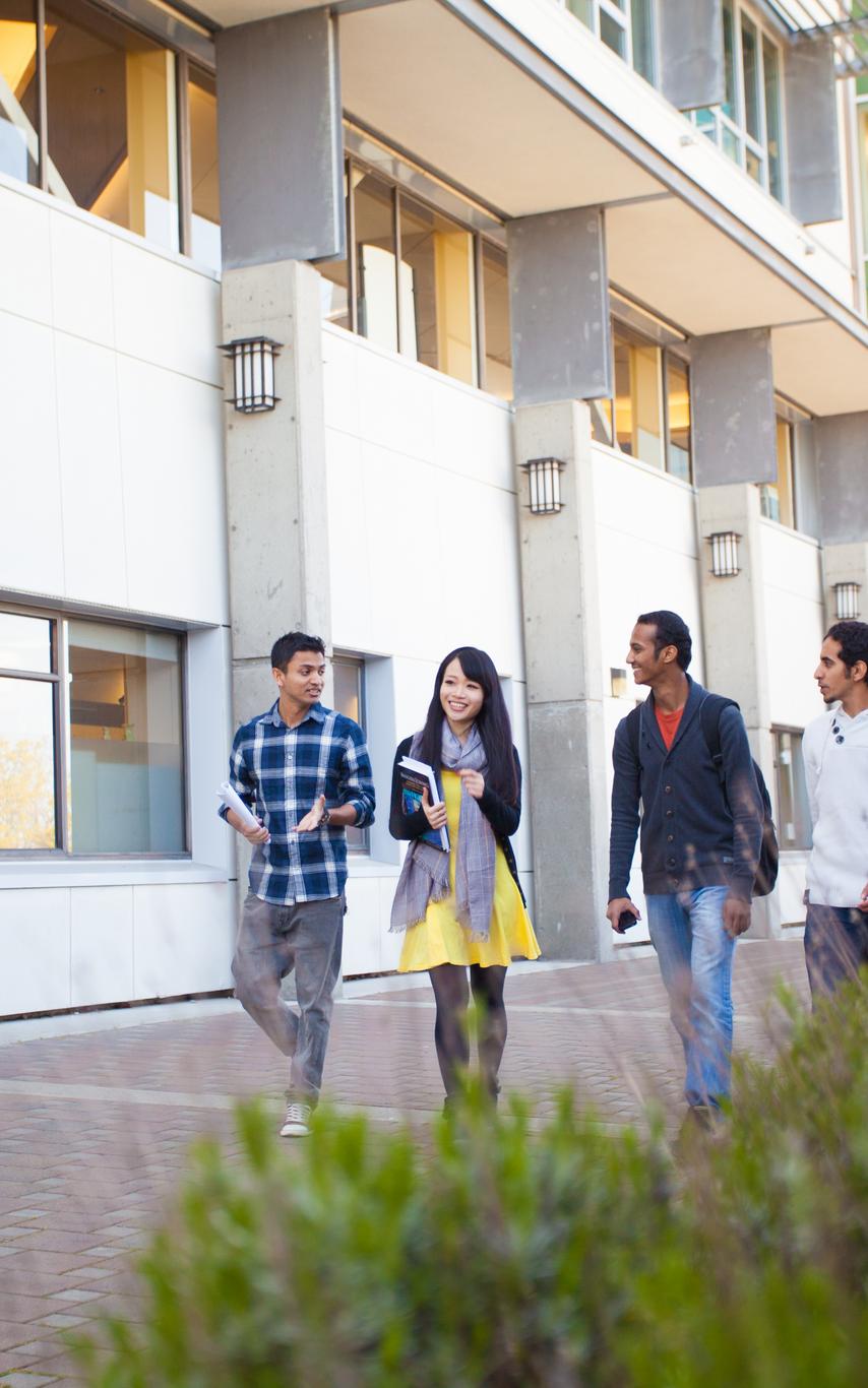 Four students walking down a path in front of a white building with windows. 