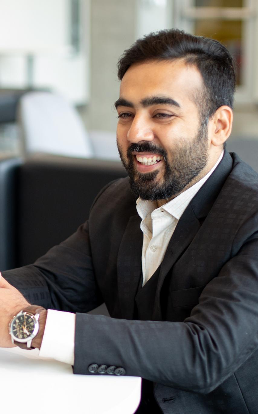 A man with short dark hair and a short beard sits at a table with his hands crossed.