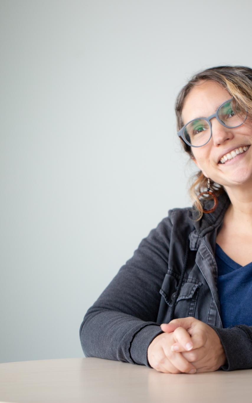 A woman smiling in grey glasses, a black jean shirt and a blue t-shirt with her arms on a table.