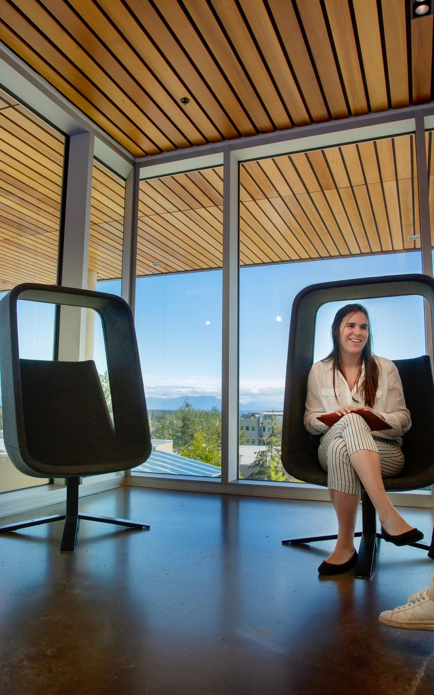 two students in grey rectangular chairs with big windows and wood slat ceiling.