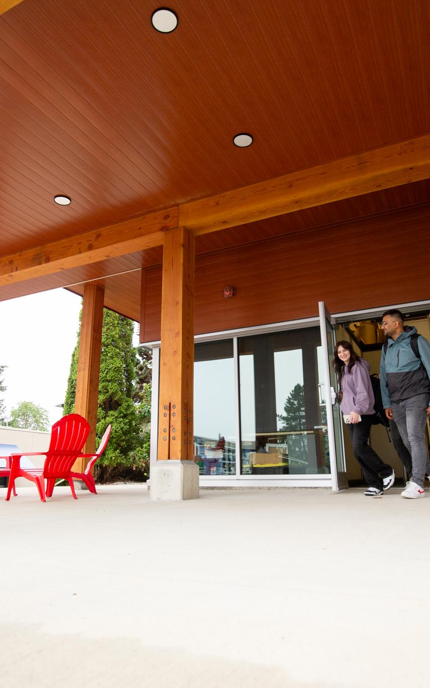 two people walking out of a a wood building with red chairs to the left of them. 