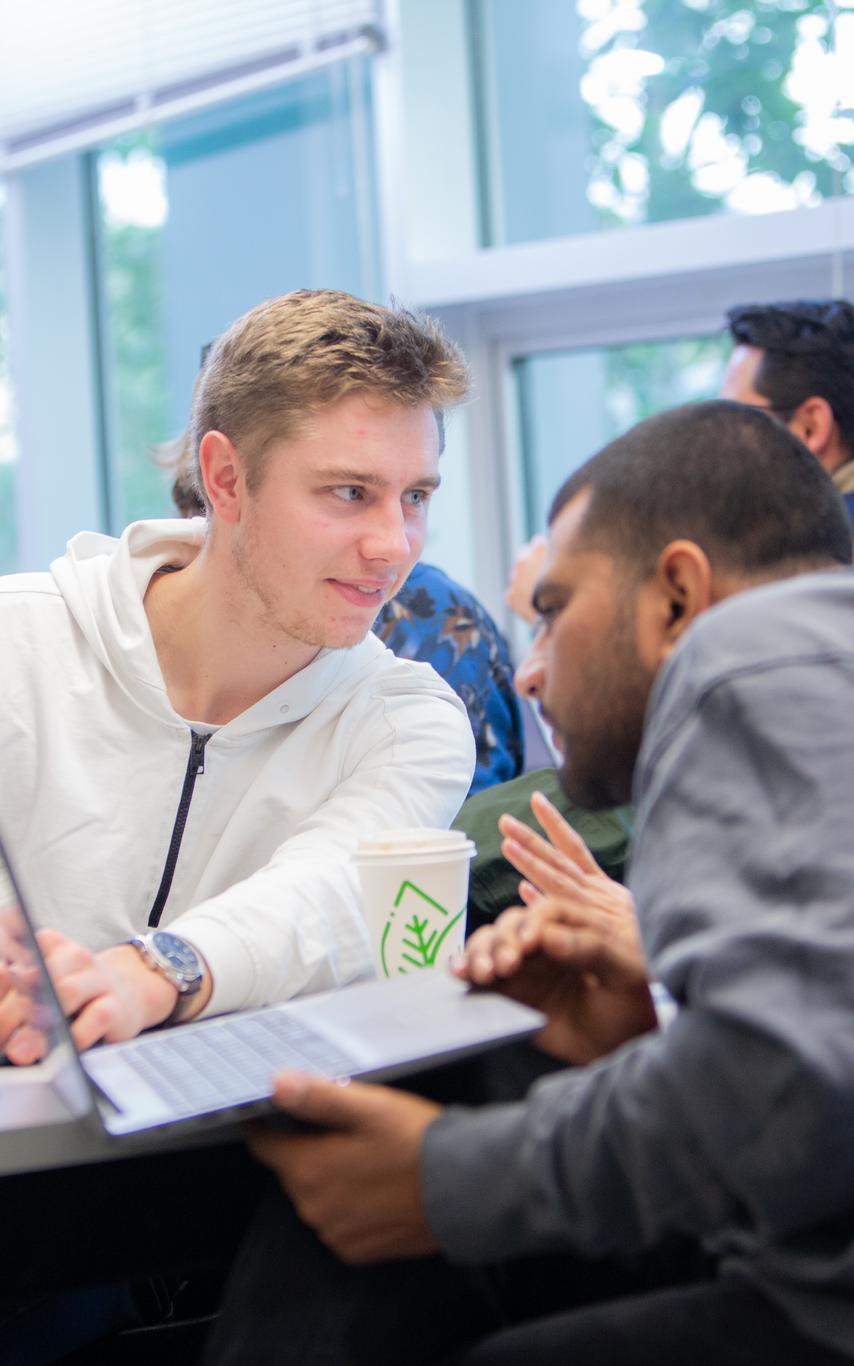 One student is looking and listening to another student while a third is looking at the computer screen, all seated at a long table 